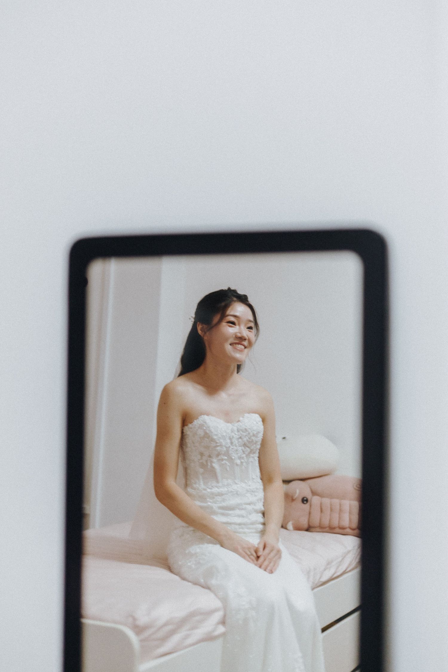 A bride in a white lace strapless wedding dress sits on a pink couch reflected in a mirror.