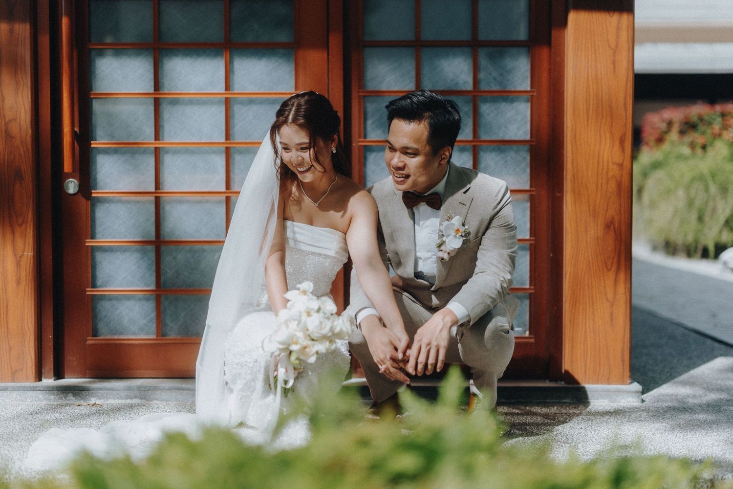 A wedding couple sits together on wooden steps outside a traditional Japanese-style building with sliding doors.