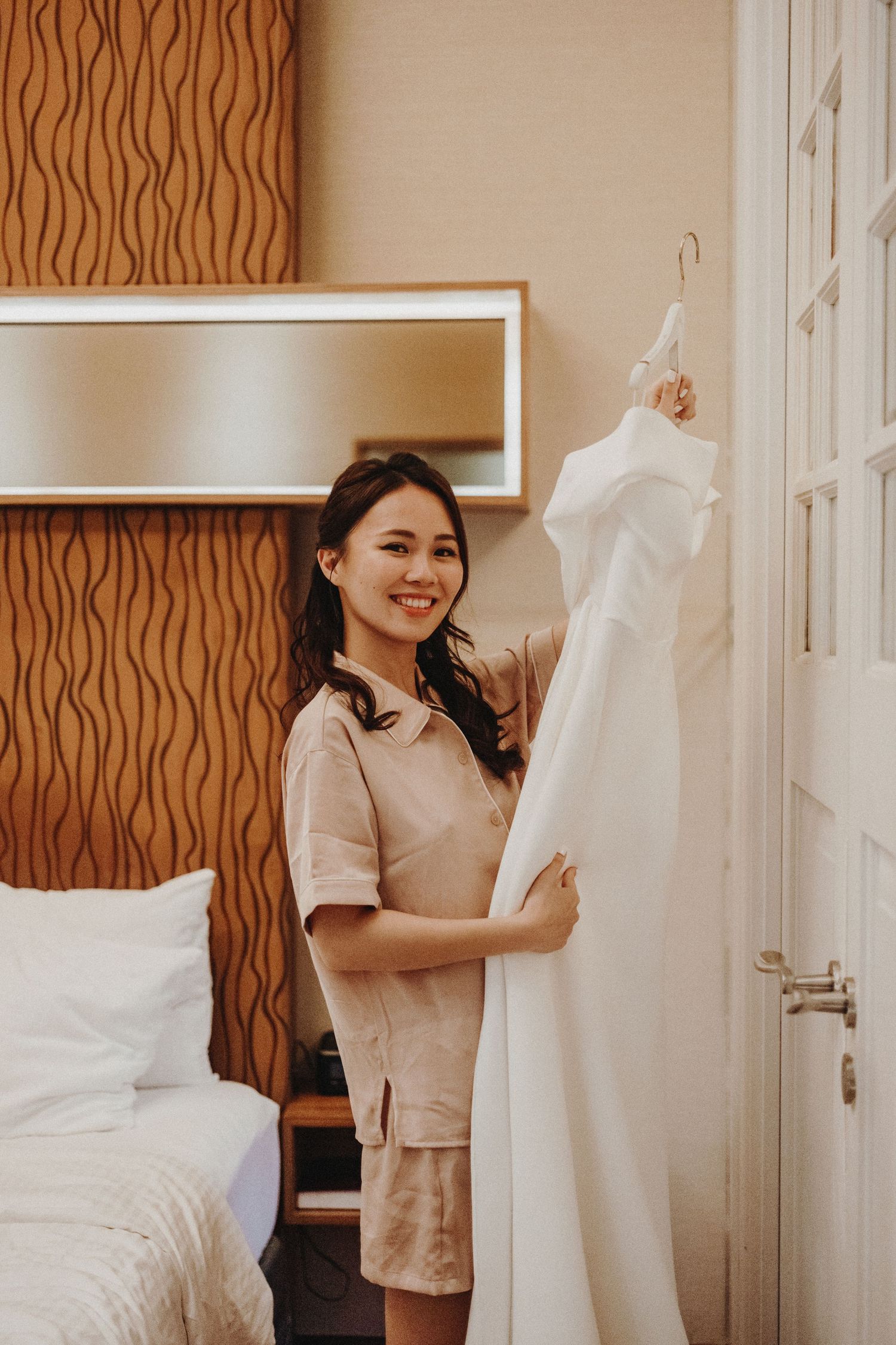 A hotel staff member in a beige uniform holds up a white wedding dress hanging near a window.