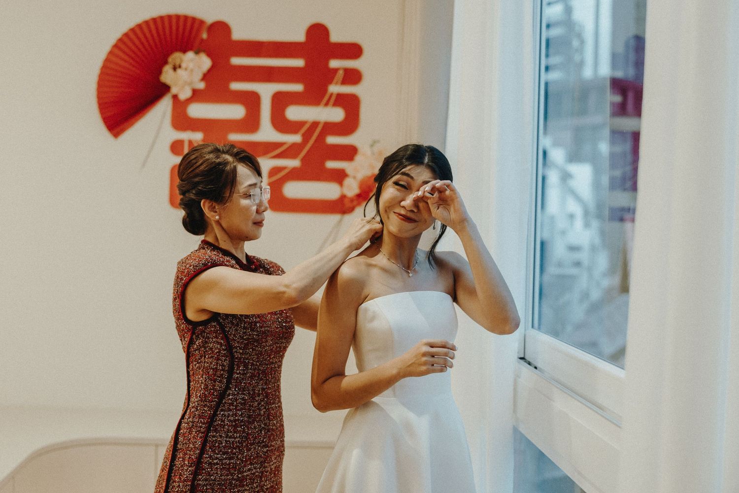 A woman in a white wedding dress stands by a window with traditional Chinese decor as someone helps with final preparations.