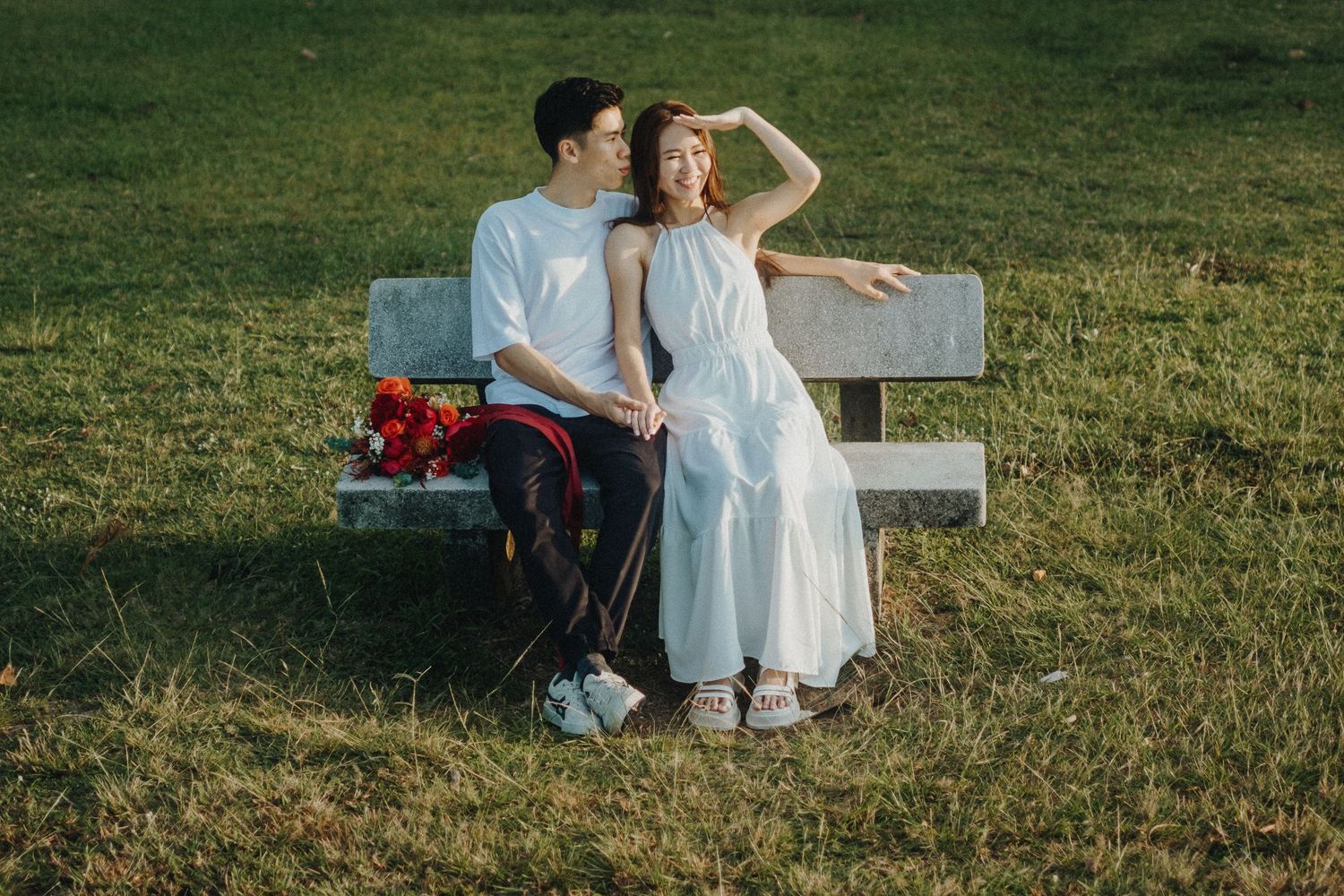 A couple sits together on a white bench in a grassy field with red flowers nearby.