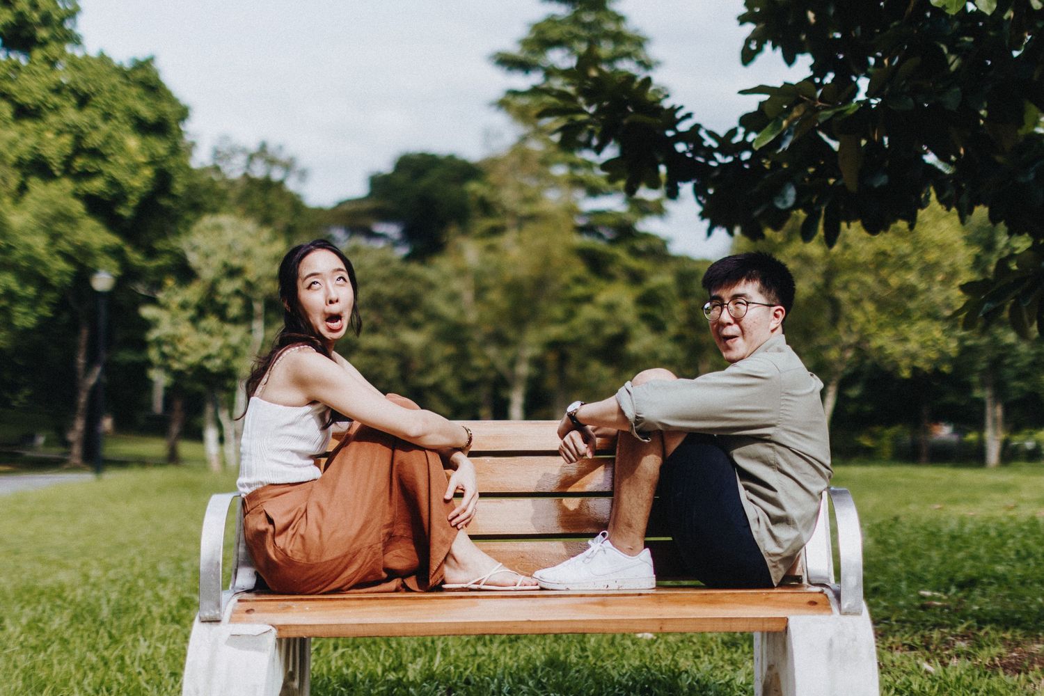 Two people sitting back-to-back on a wooden park bench surrounded by green trees.