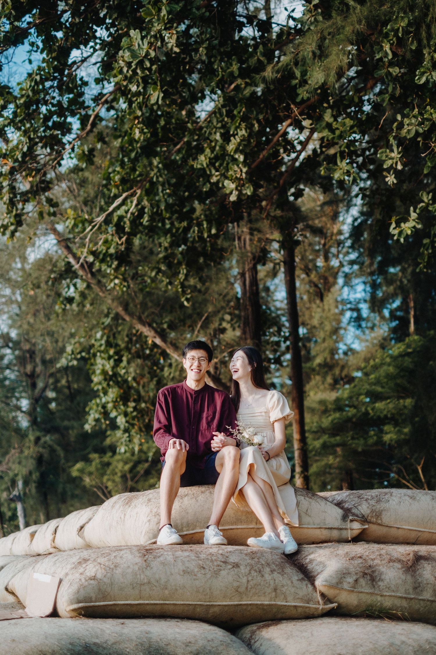 A couple sits together on sandbags under green trees on a sunny afternoon.