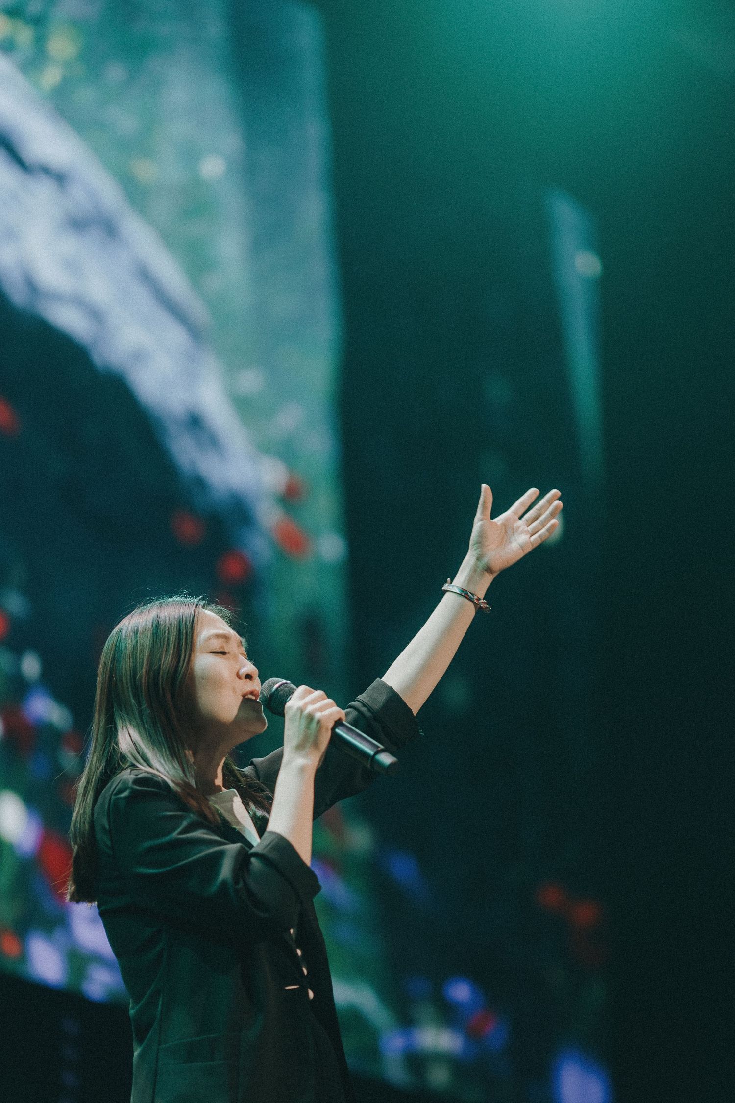 Singer in a black outfit performs passionately on stage with raised arm under dramatic lighting and effects.
