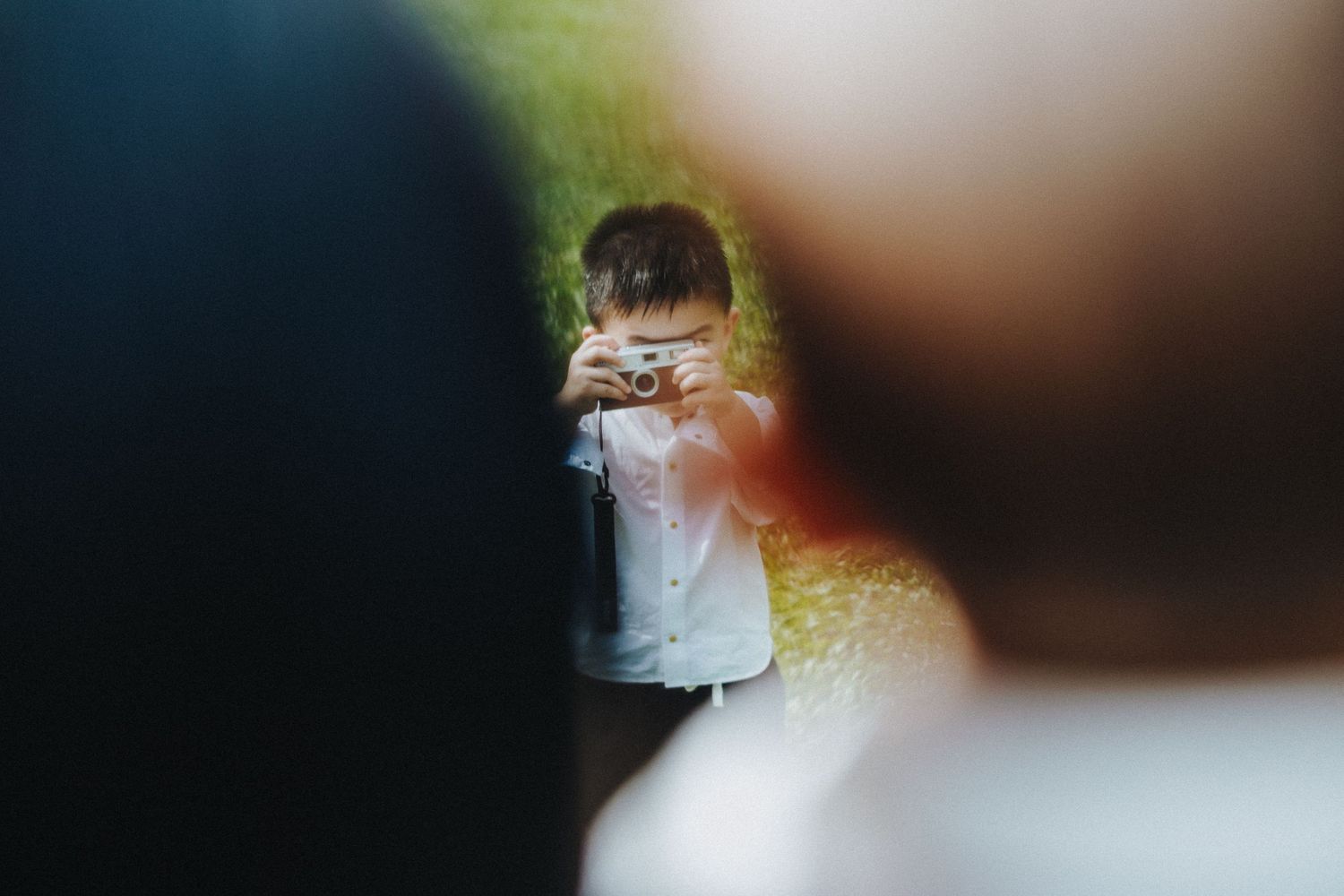 A child holds up a camera to take a photo while wearing a white shirt in a bokeh-filled outdoor setting.