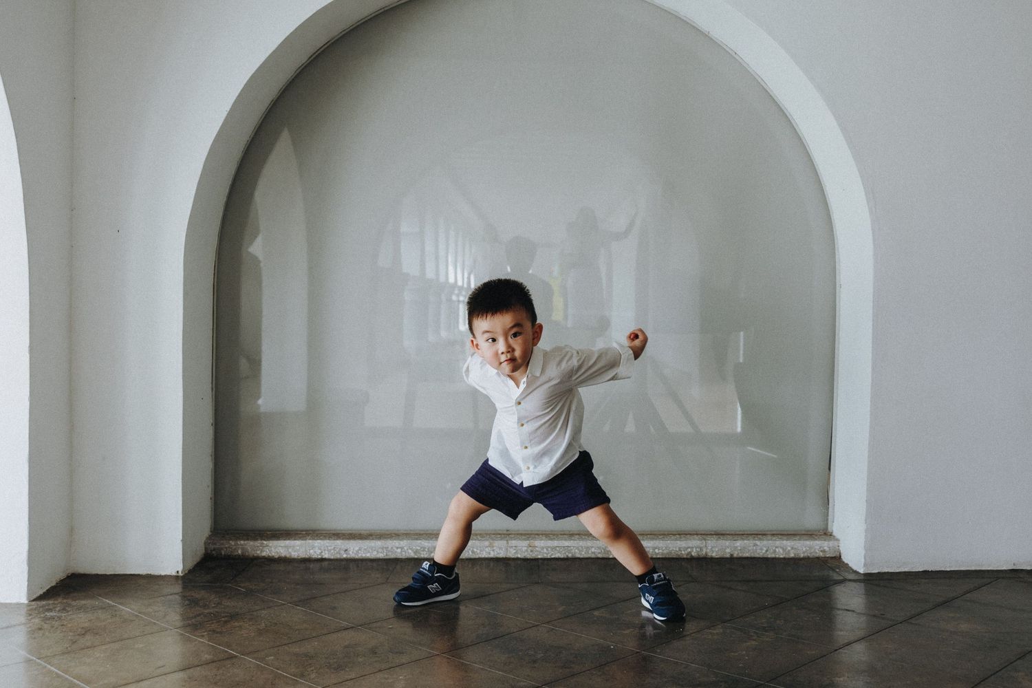 A child in a white shirt and blue shorts performs an energetic jumping pose in a bright minimalist space.