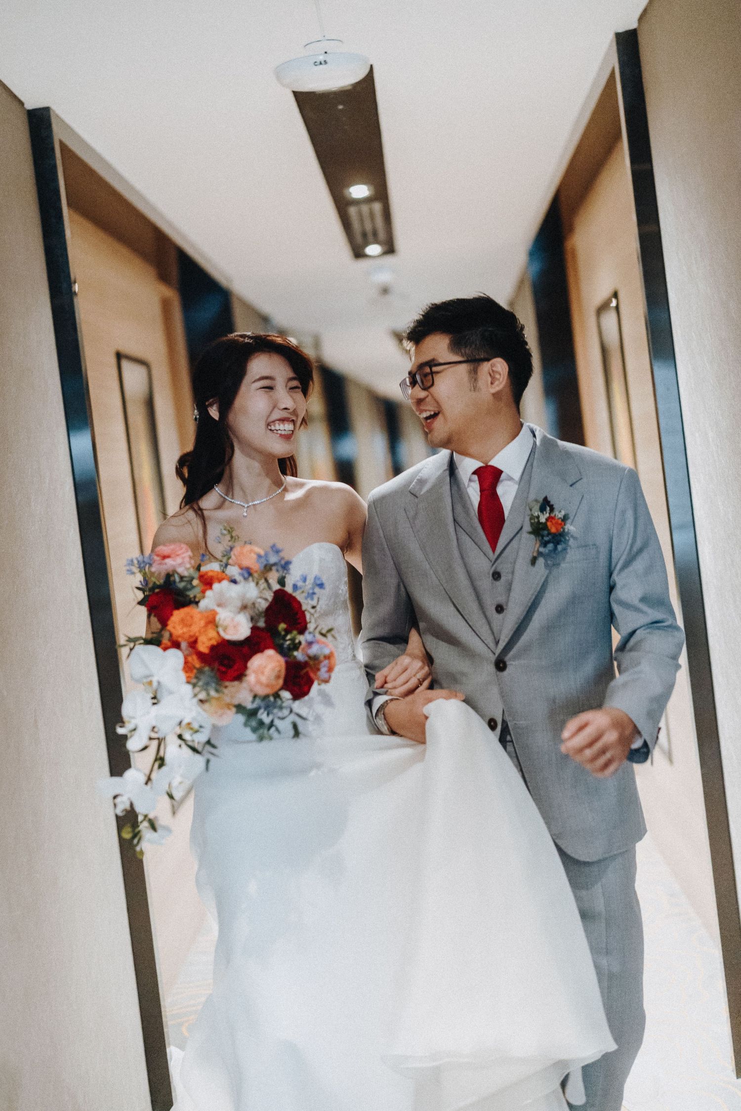 A couple share joyful moments in a hotel hallway during their wedding photoshoot wearing grey suit and white gown.
