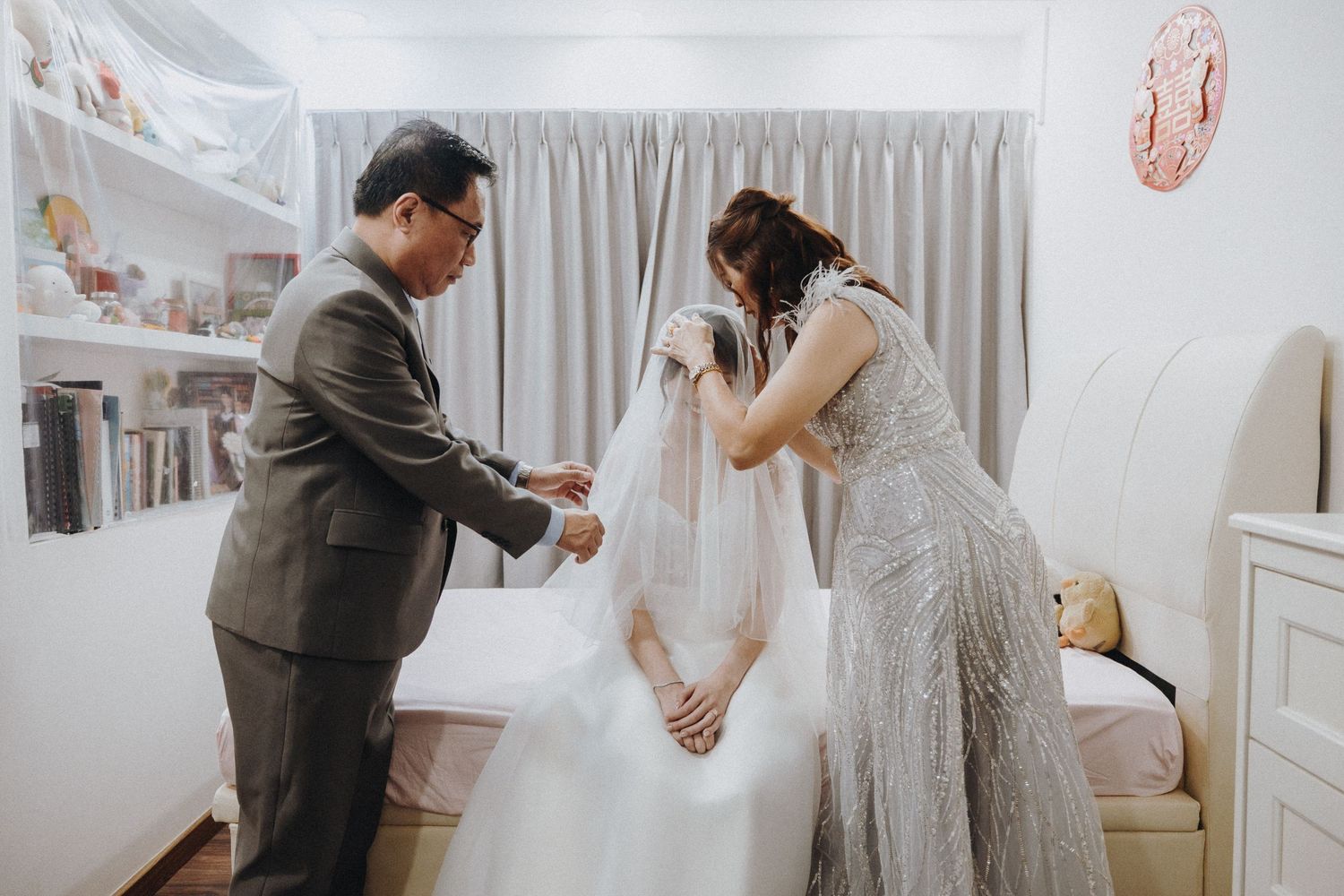 Two people help adjust a feminine white wedding dress in a softly lit bedroom setting.