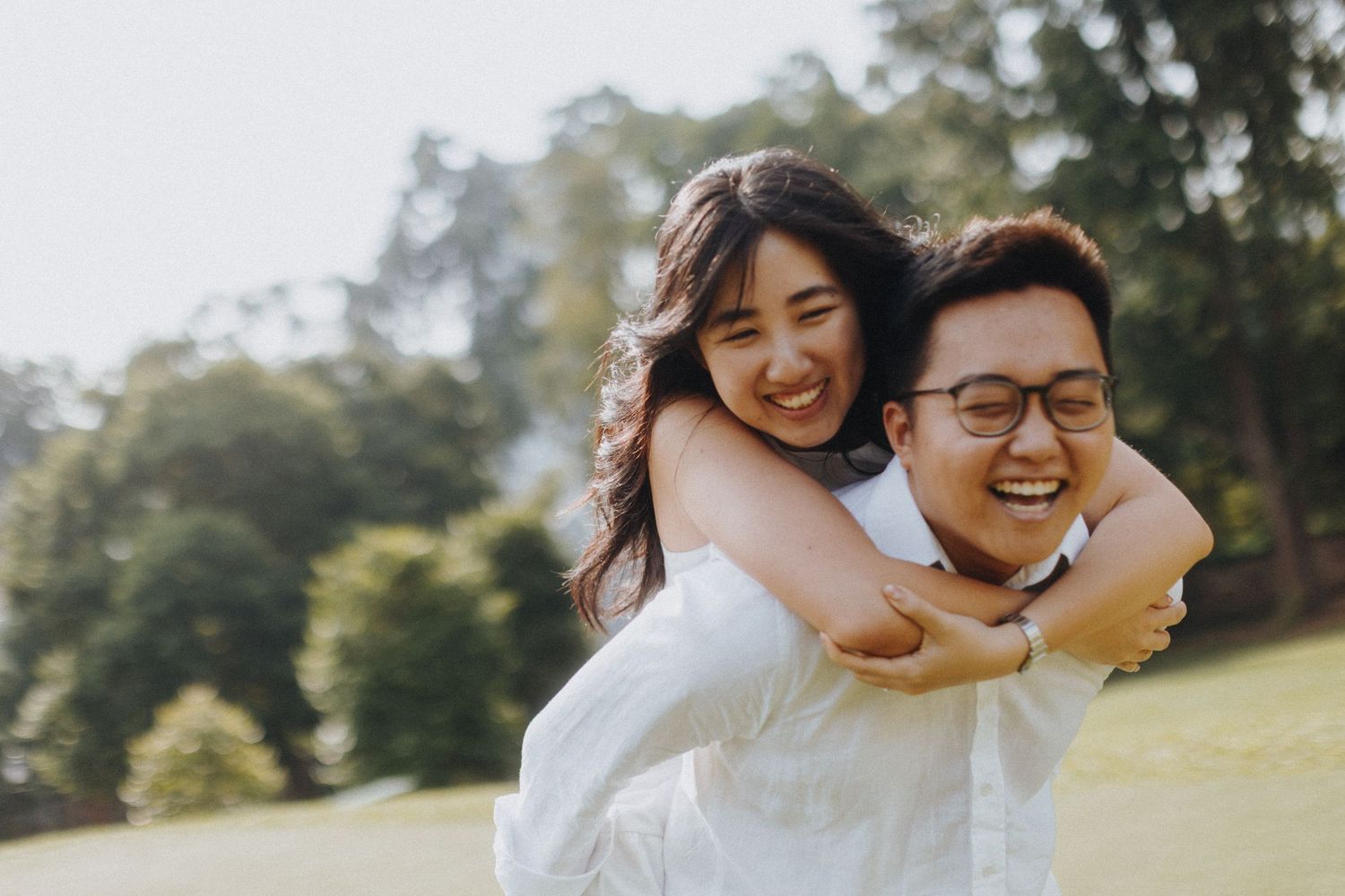 A couple shares a piggyback ride moment outdoors in a sunny park setting.