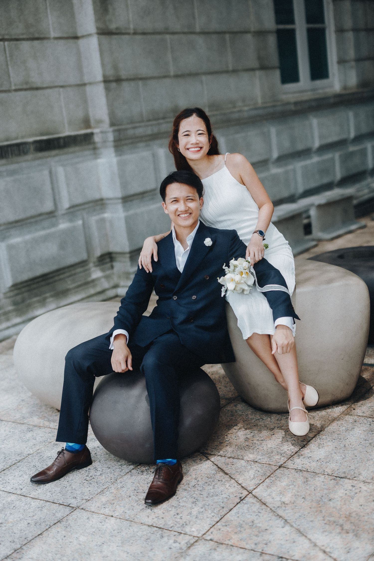 Couple sits casually on grey bean bag chairs against a concrete wall in an outdoor setting.