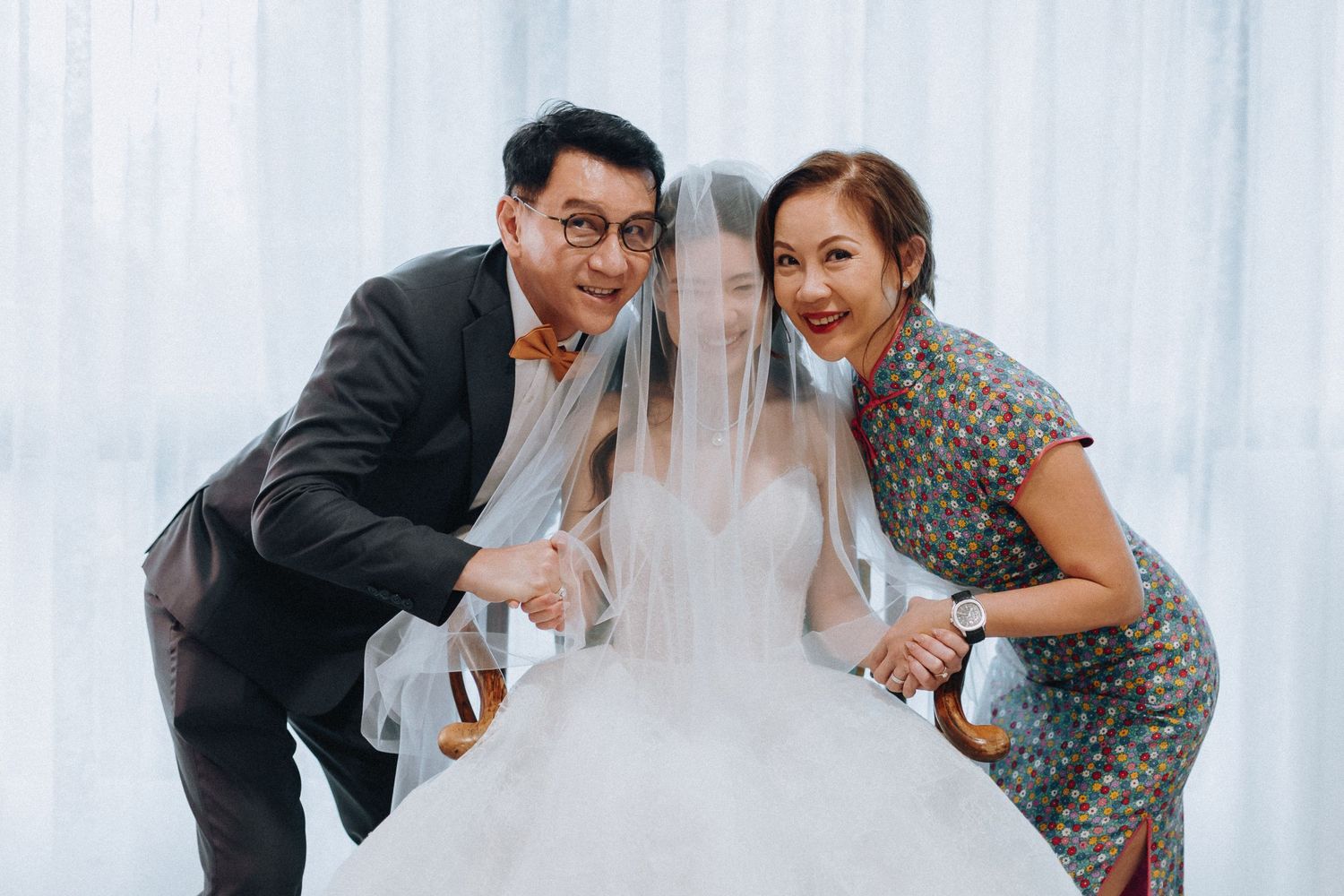 Happy parents posing with veiled bride in white wedding gown during pre-wedding preparations.