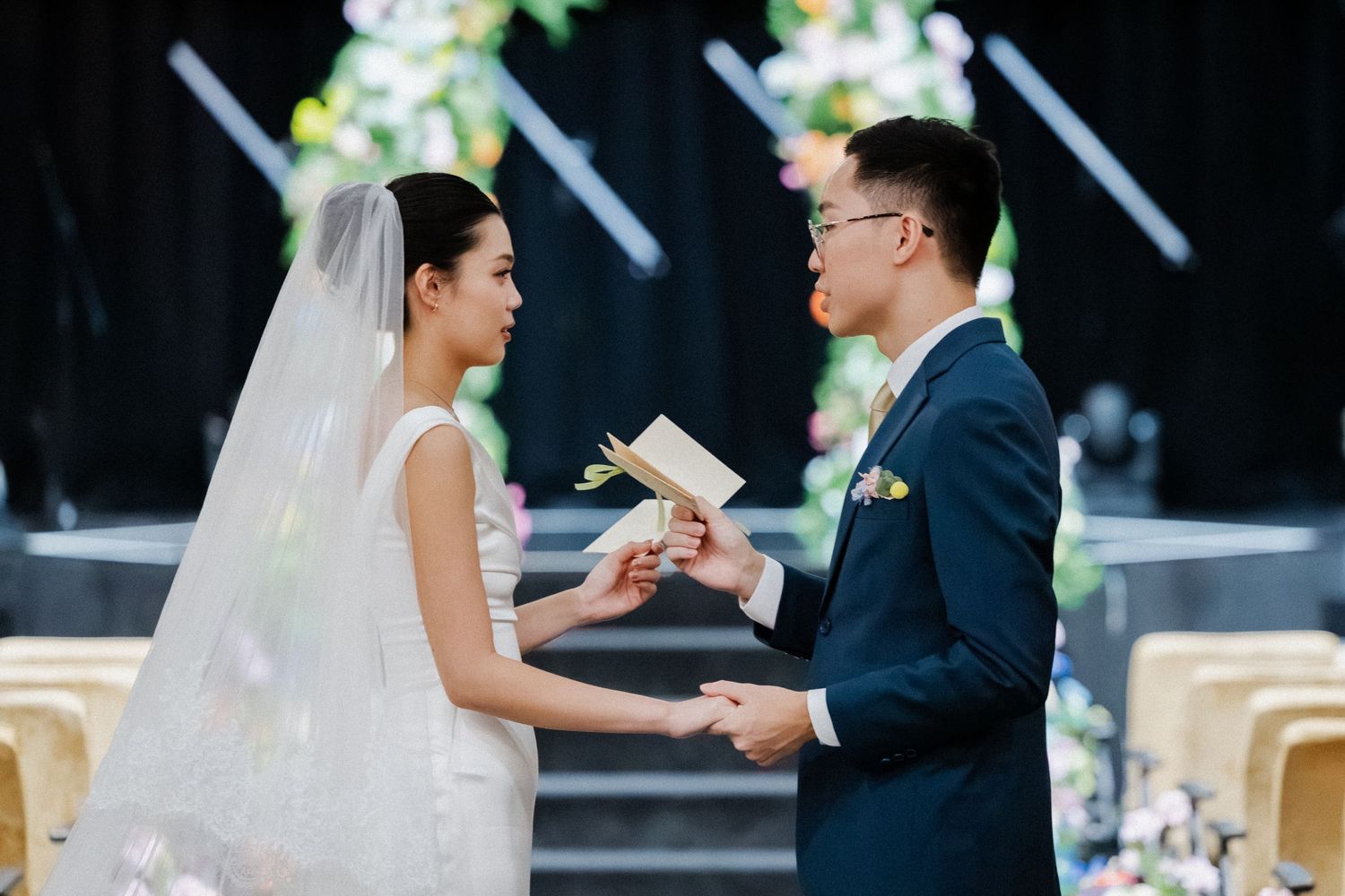 Asian bride and groom exchanging vows at wedding altar, groom reading from card while holding bride's hands at floral ceremony.
