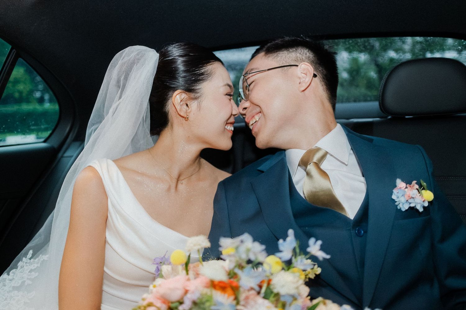 Romantic bride and groom share a tender nose-to-nose moment in car backseat with colorful wedding bouquet.