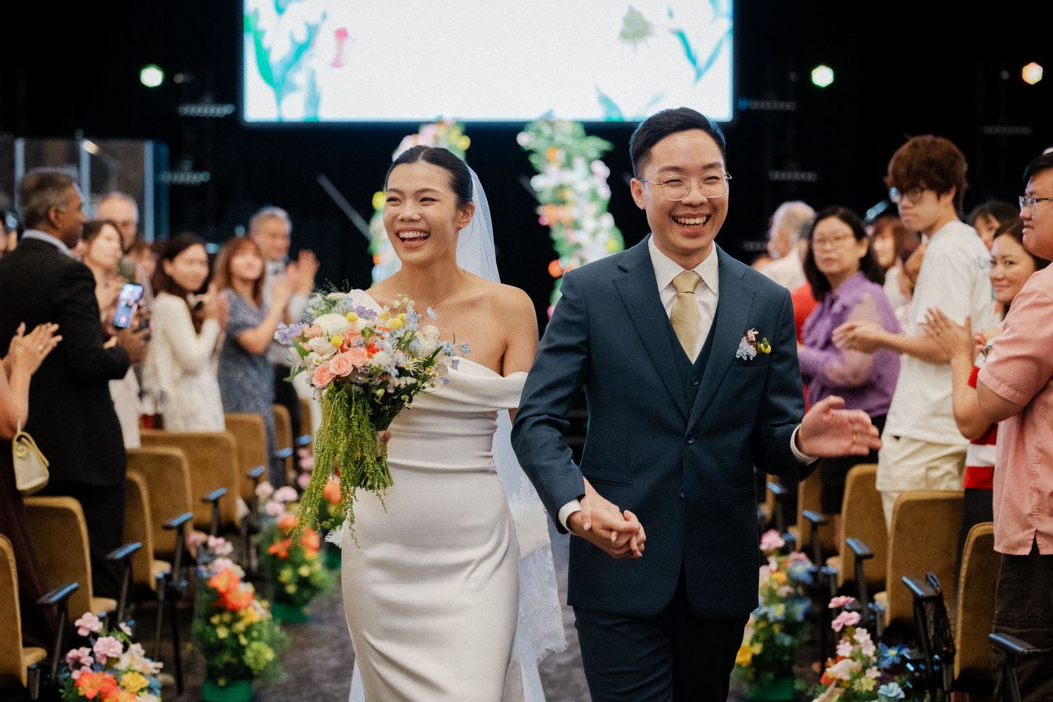 Happy couple holding hands walking down floral-lined aisle after wedding ceremony.