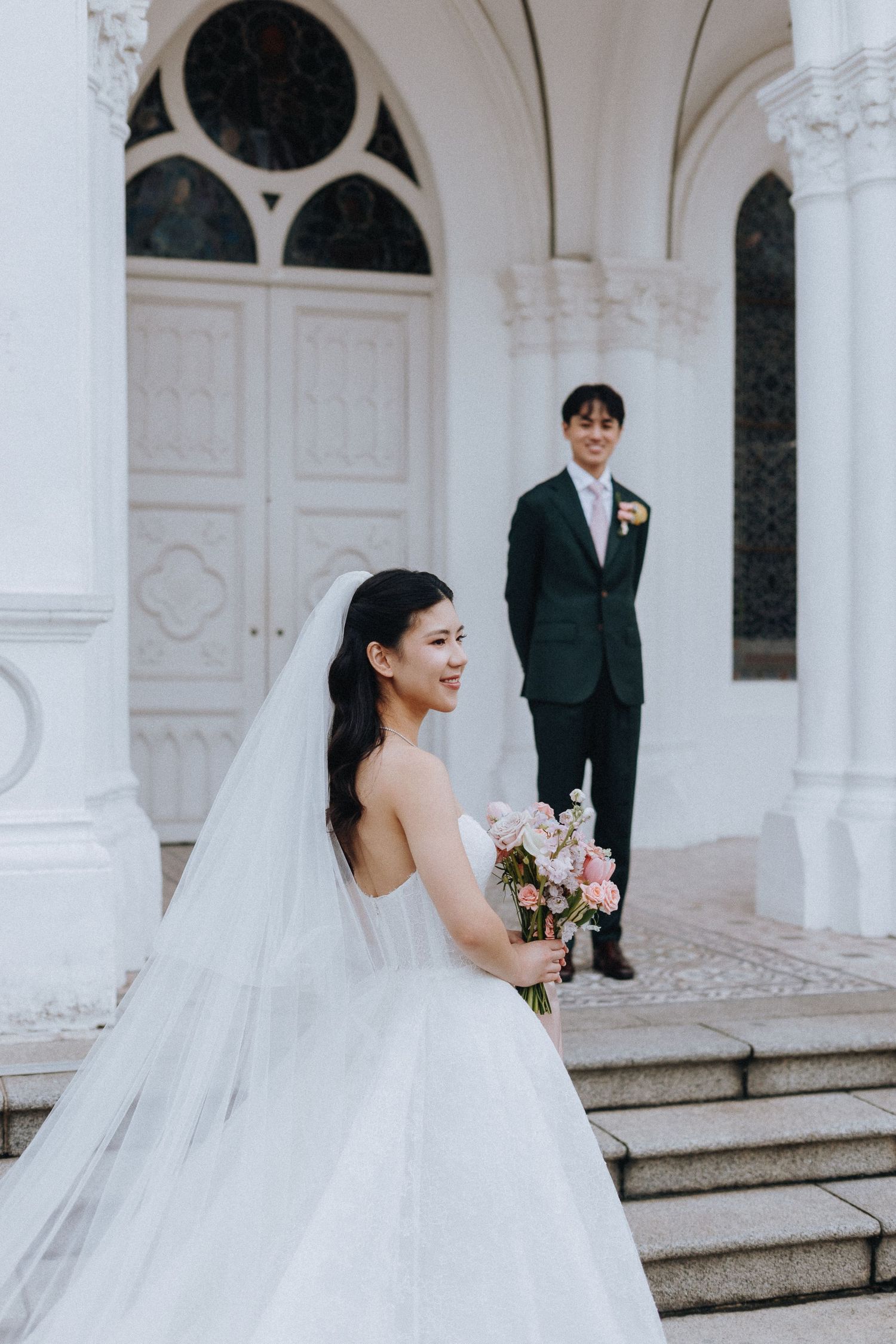 Asian bride in a white gown holding pink flowers stands before a groom at a white church entrance.