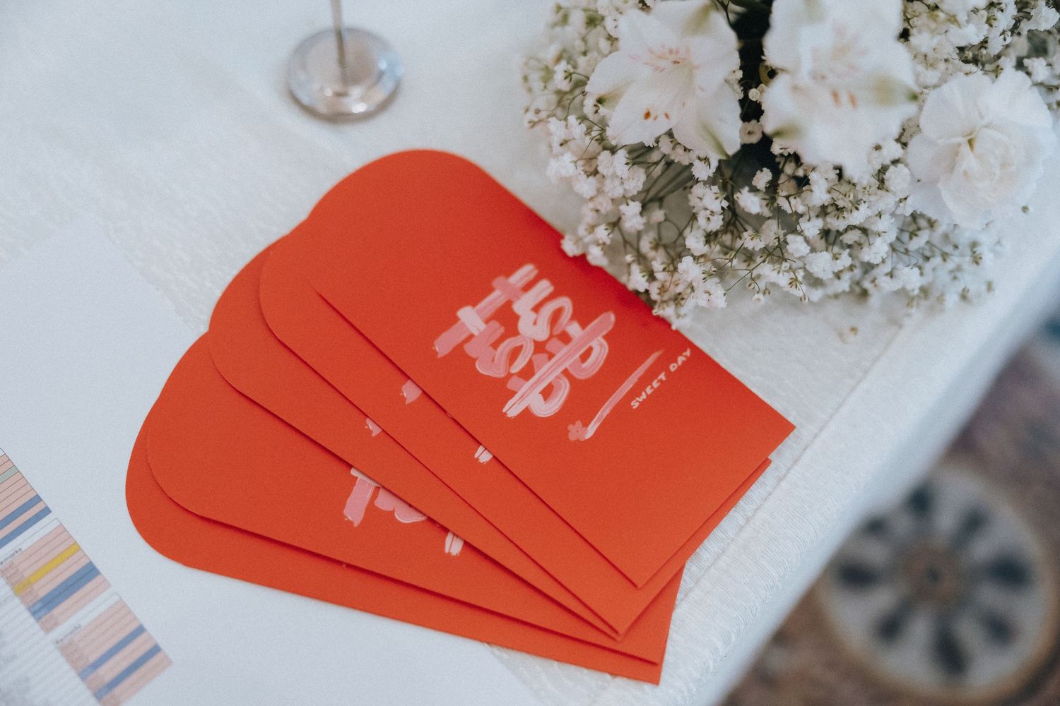 Red Chinese double happiness envelopes displayed on a white table next to white flowers at a wedding reception.