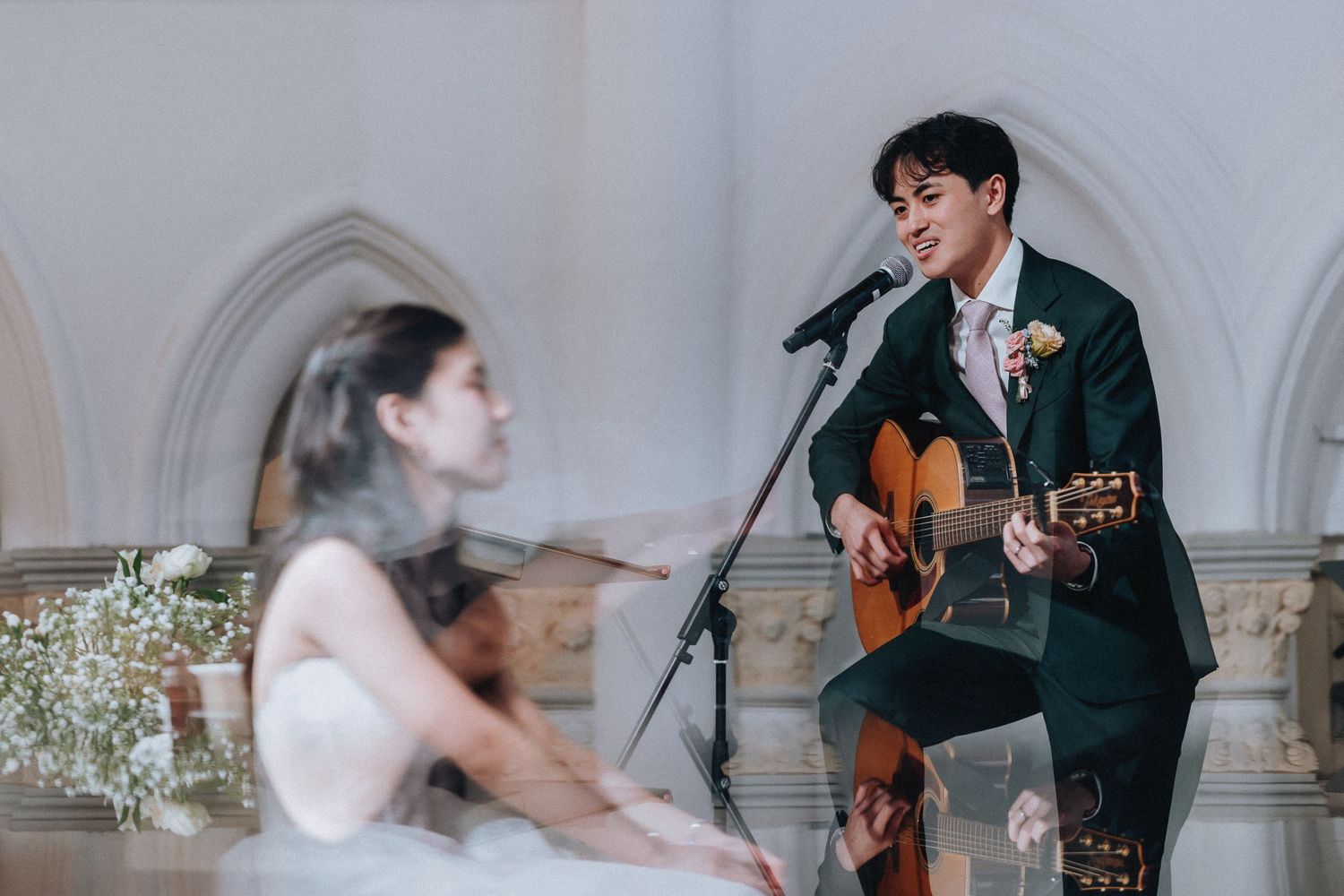 Musician plays guitar and sings at a wedding while bride plays violin on stage.