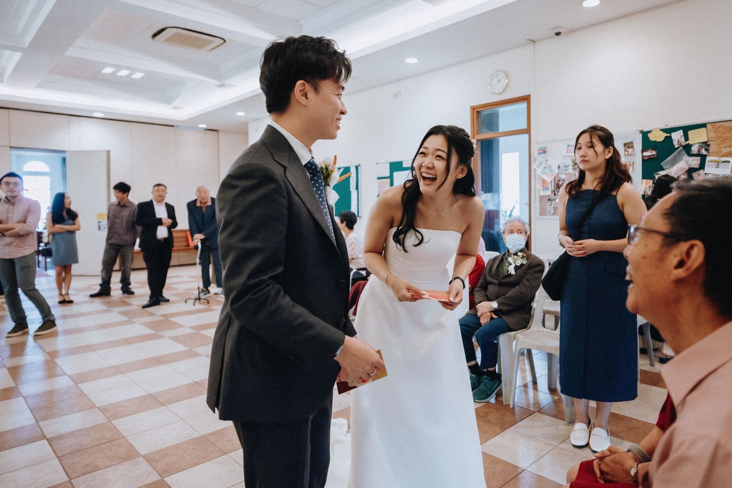 Bride laughs joyfully with groom and guests in bright hall during wedding reception celebration.