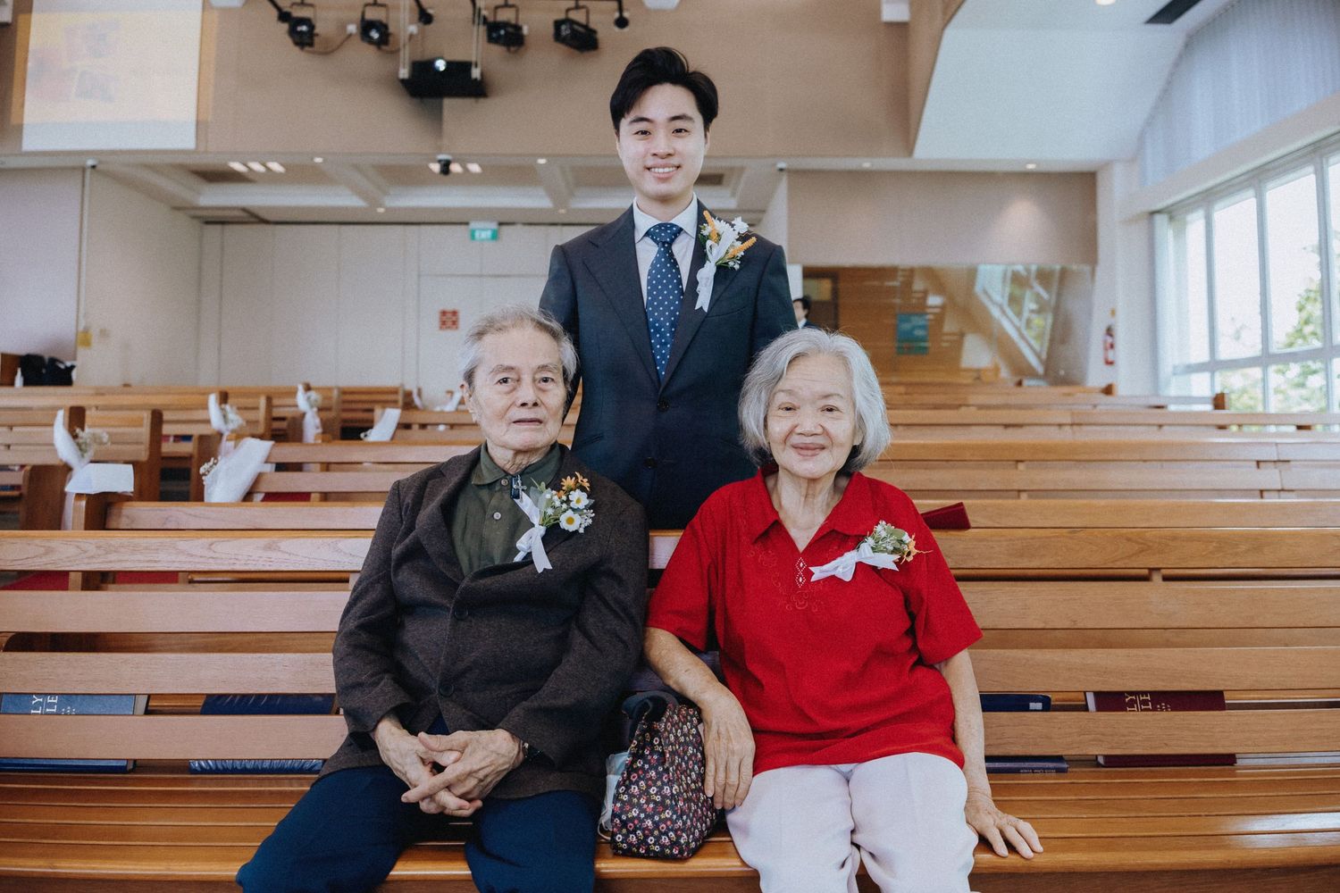 Groom poses with elderly grandparents seated on church pew before wedding ceremony.
