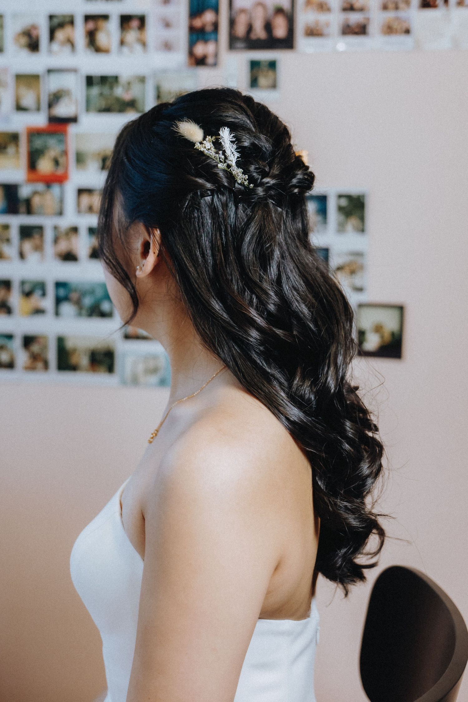 An elegant bridal half-up hairstyle adorned with delicate dried flowers against a wall of polaroid photos.
