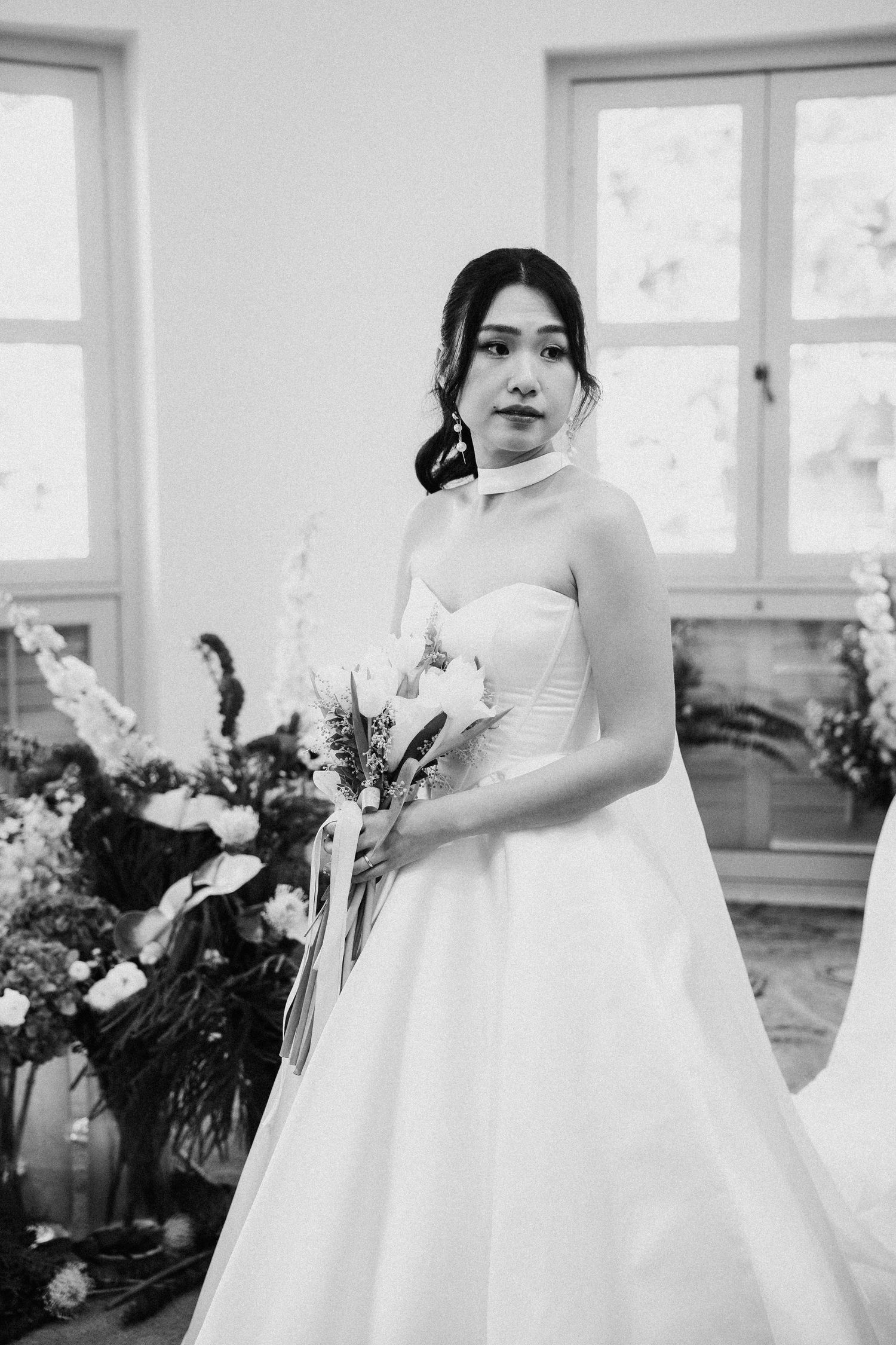 Black and white photo of a bride in a strapless ball gown holding a bouquet, standing before floral arrangements.