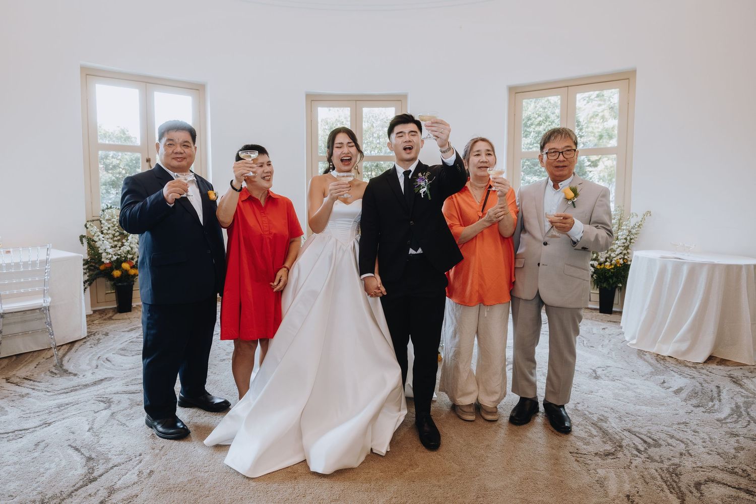 Wedding couple celebrates with four family members raising glasses in bright white reception venue.