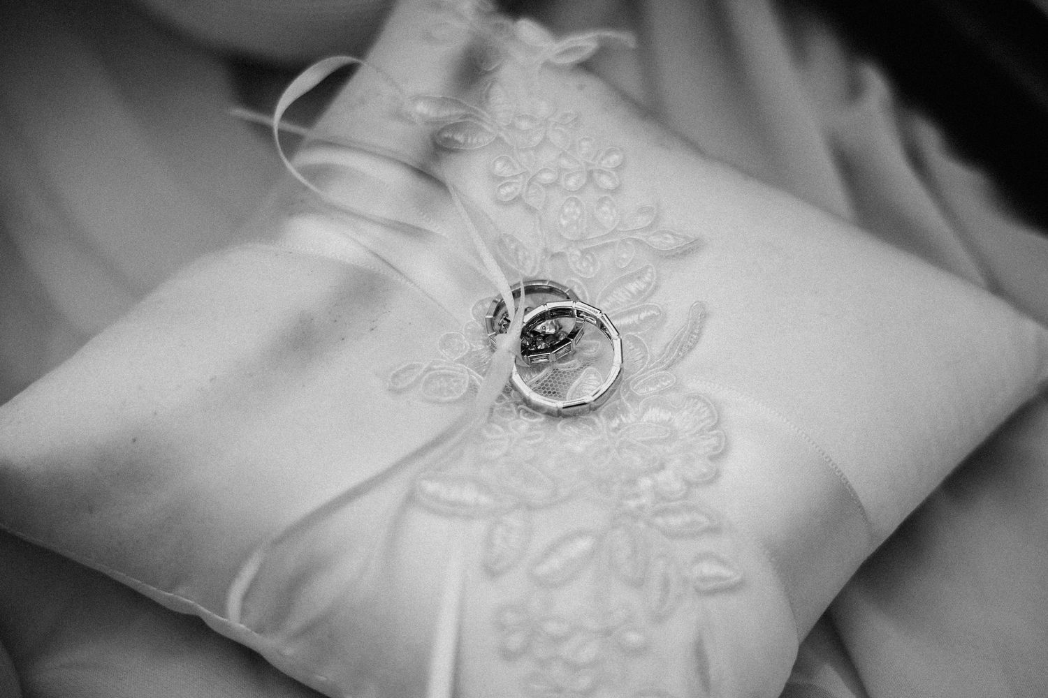 Two wedding rings resting on an embroidered white satin ring pillow in a black and white photo.