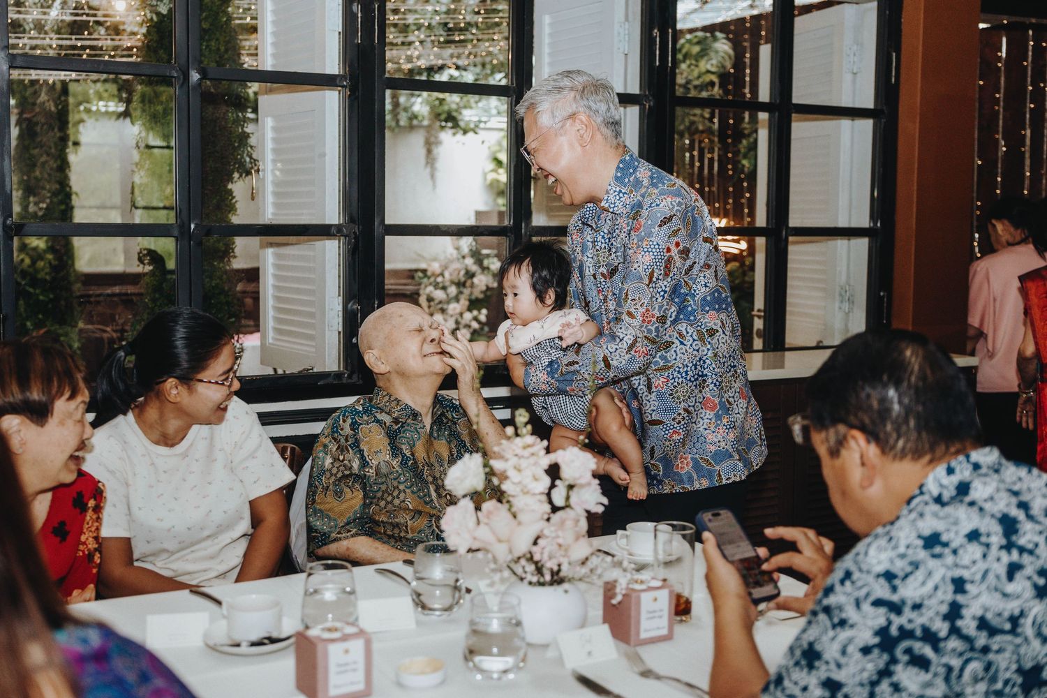 An elderly man holds a baby while greeting seated guests at a wedding reception table.
