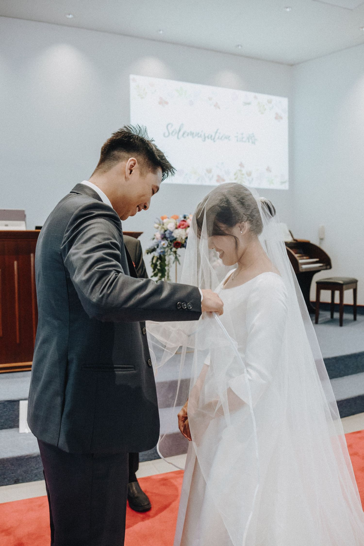 Groom lifting bride's veil during wedding ceremony in a church with a solemnization sign in the background.