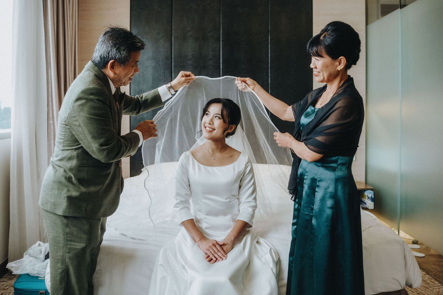 Parents helping bride with veil before wedding ceremony in hotel room.