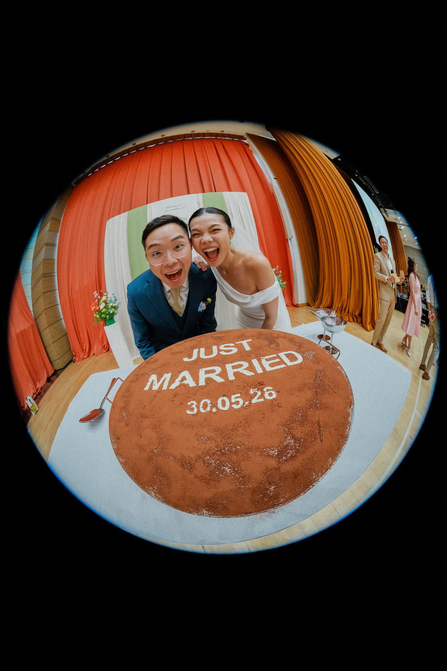 Newlywed couple poses joyfully behind a round wedding cake reading Just Married 30.05.26 in a fisheye lens photo.