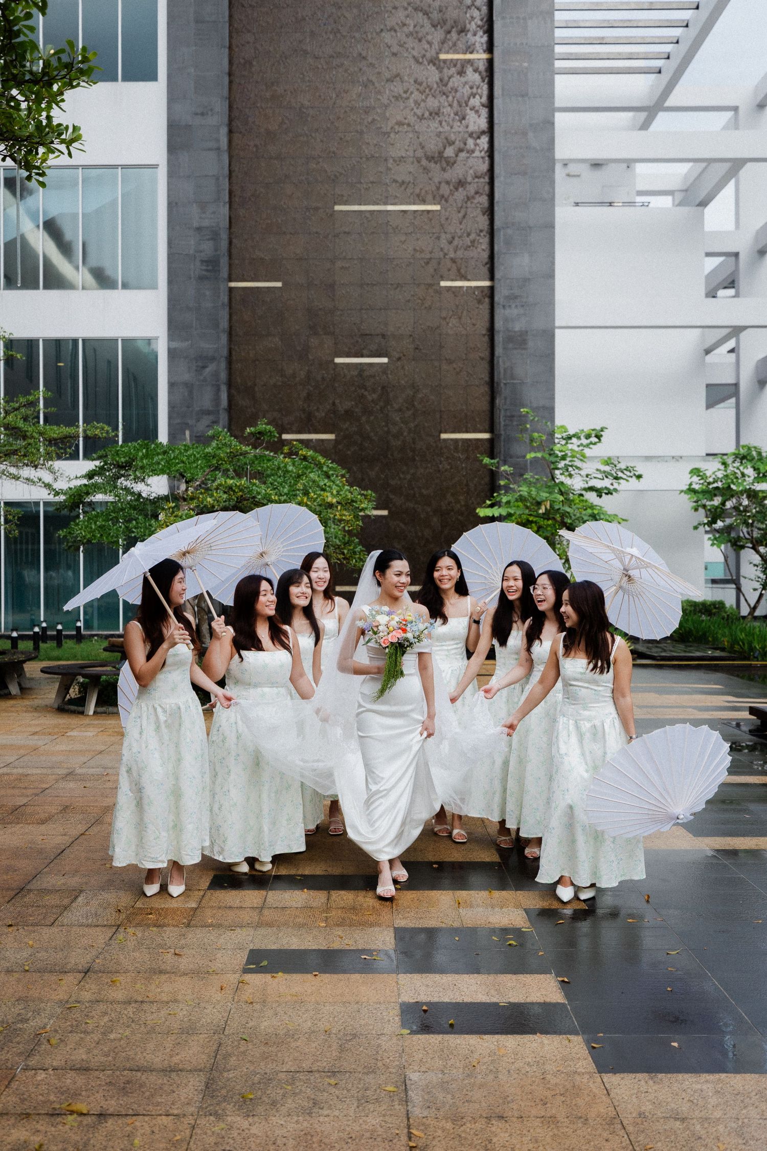 Bride and bridesmaids in white dresses holding parasols pose together outdoors at a modern wedding venue.