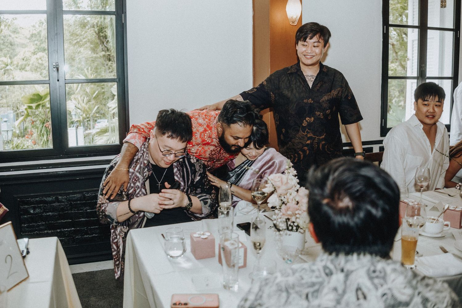 Wedding guests laugh and gather around table during lively indoor reception celebration.
