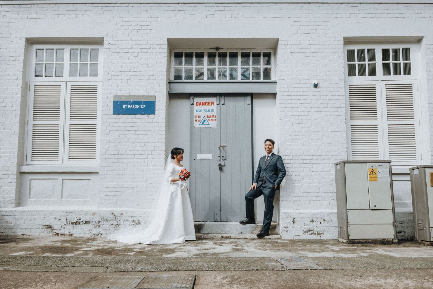 Bride and groom pose outside a white brick building with a grey door on their wedding day.