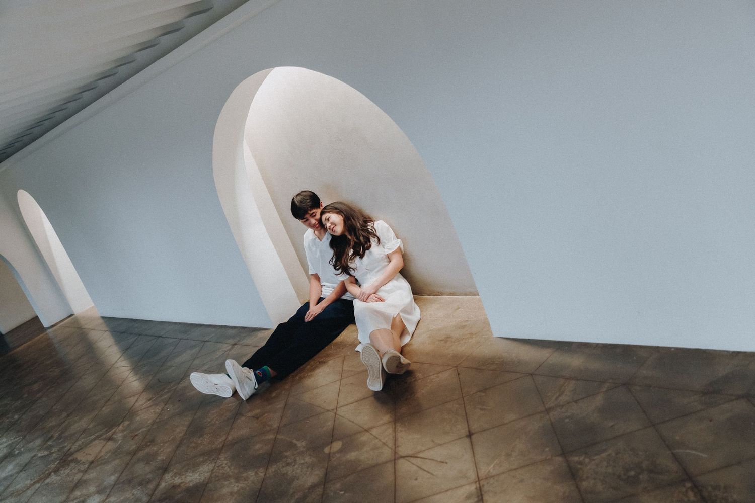 Couple sits intimately together in a minimalist architectural space with white arched walls and wooden flooring.