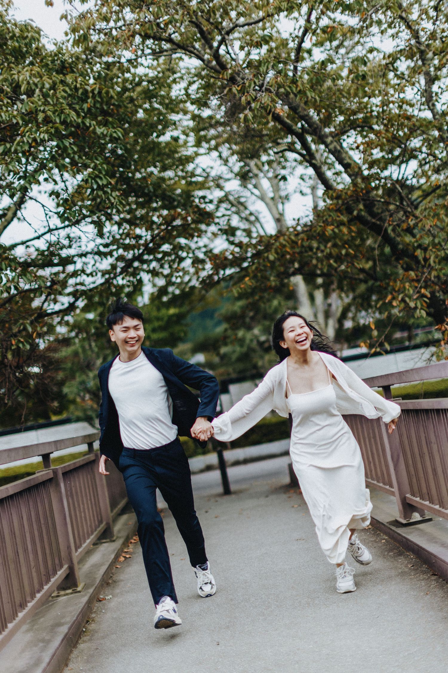 A joyful couple in casual and formal attire running together on a wooden bridge path surrounded by green trees.