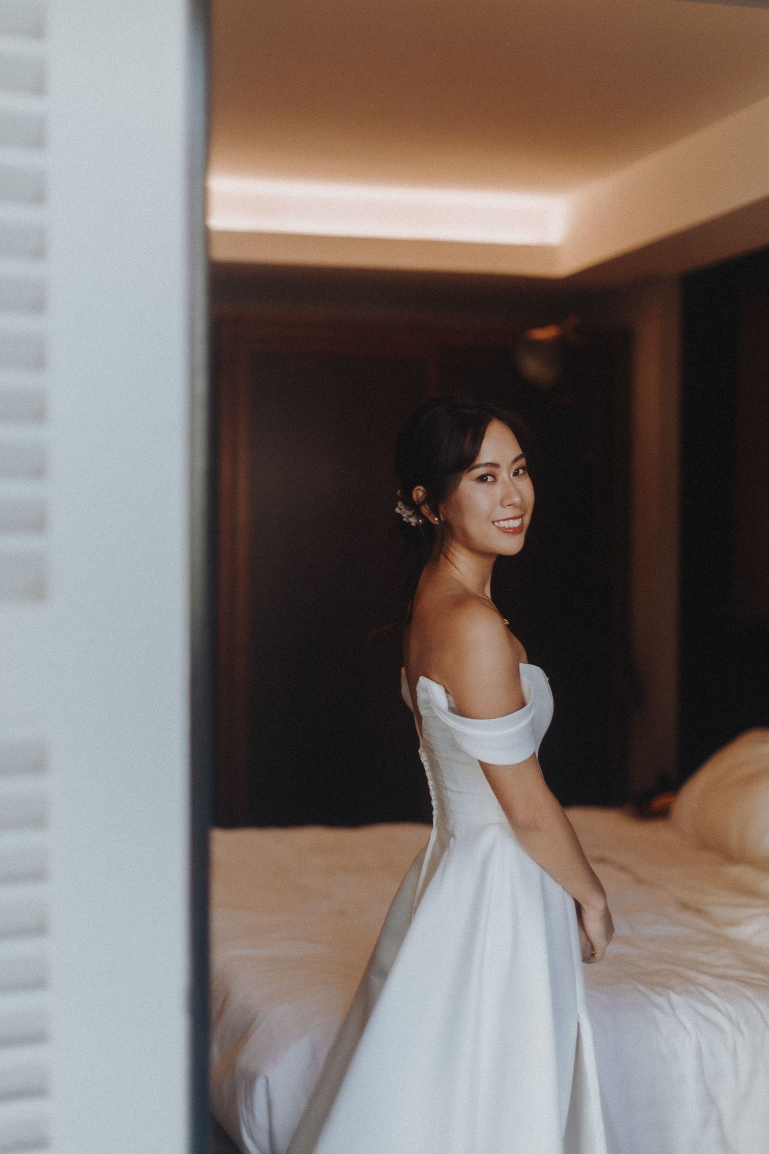 A bride in an elegant off-shoulder white wedding gown smiles while getting ready in a hotel room.