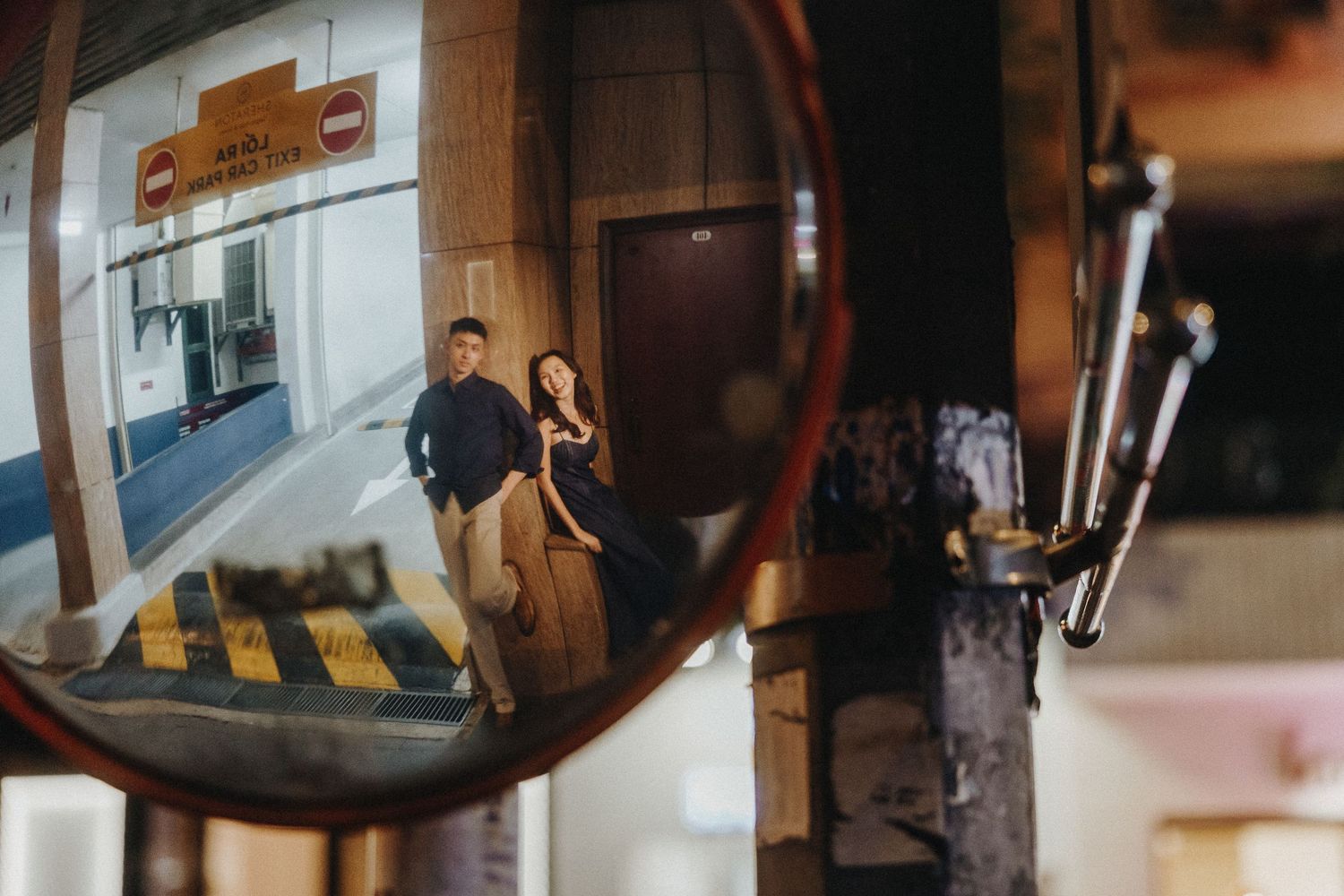 A convex security mirror at night reflects figures walking past a brightly lit storefront entrance.