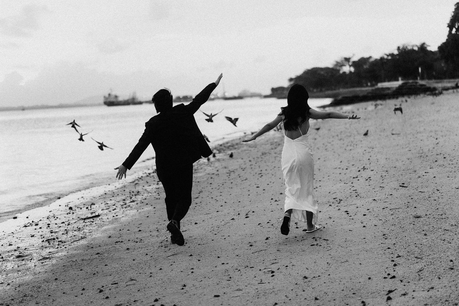 Playful couple running along the shoreline with dramatic black and white contrast.