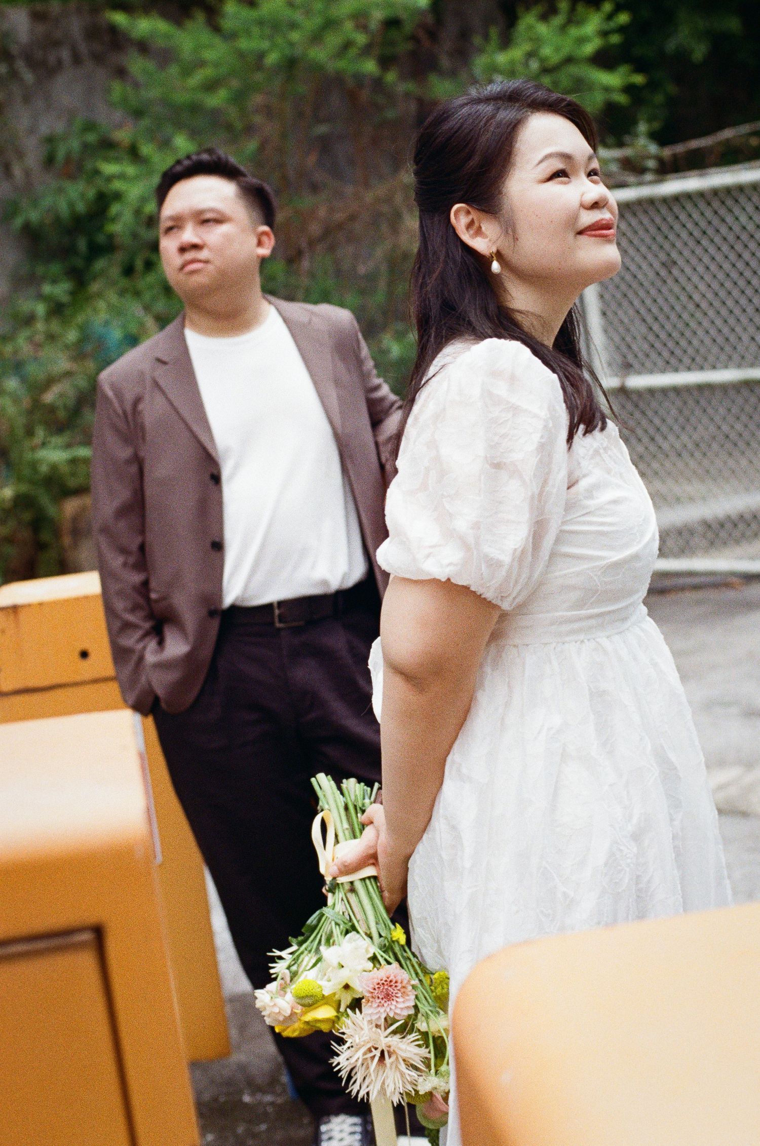 A couple poses outdoors, with the woman in a white dress holding a mixed floral bouquet.