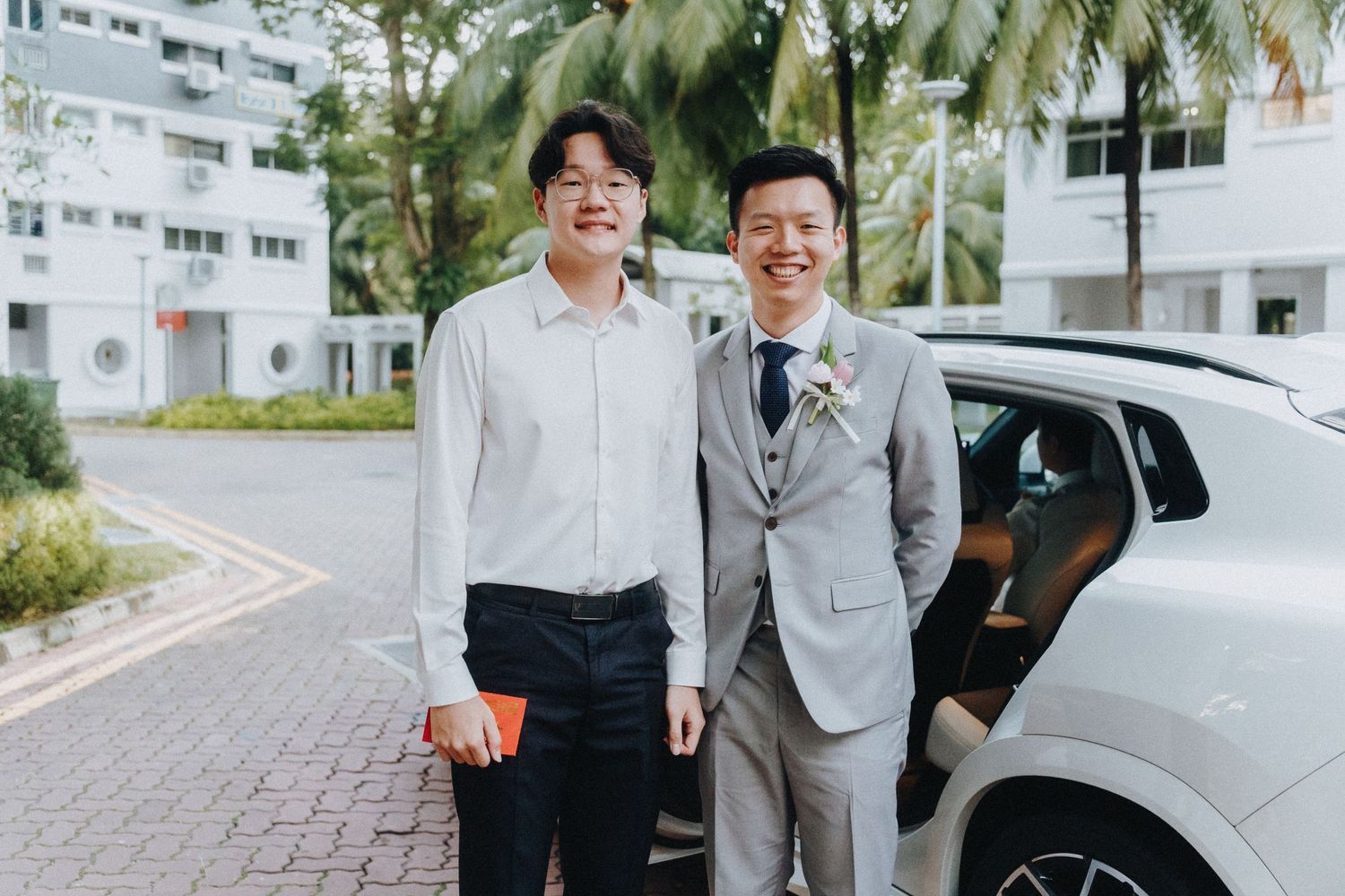 Two people in formal attire standing and smiling next to a parked car on a palm tree-lined street.