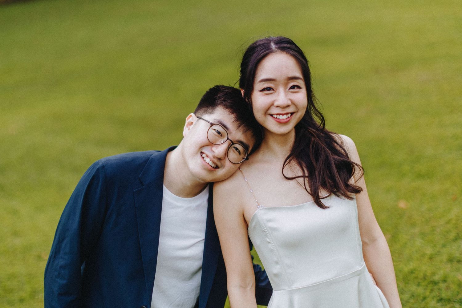 A couple in casual wedding attire pose together outdoors on a grassy lawn on a sunny day.