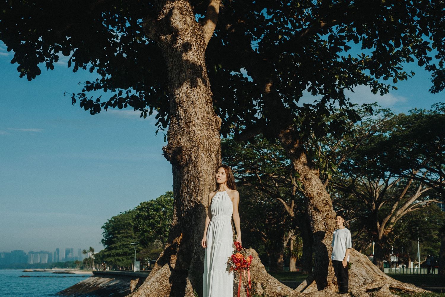 Woman in white dress poses by large tree with ocean view.