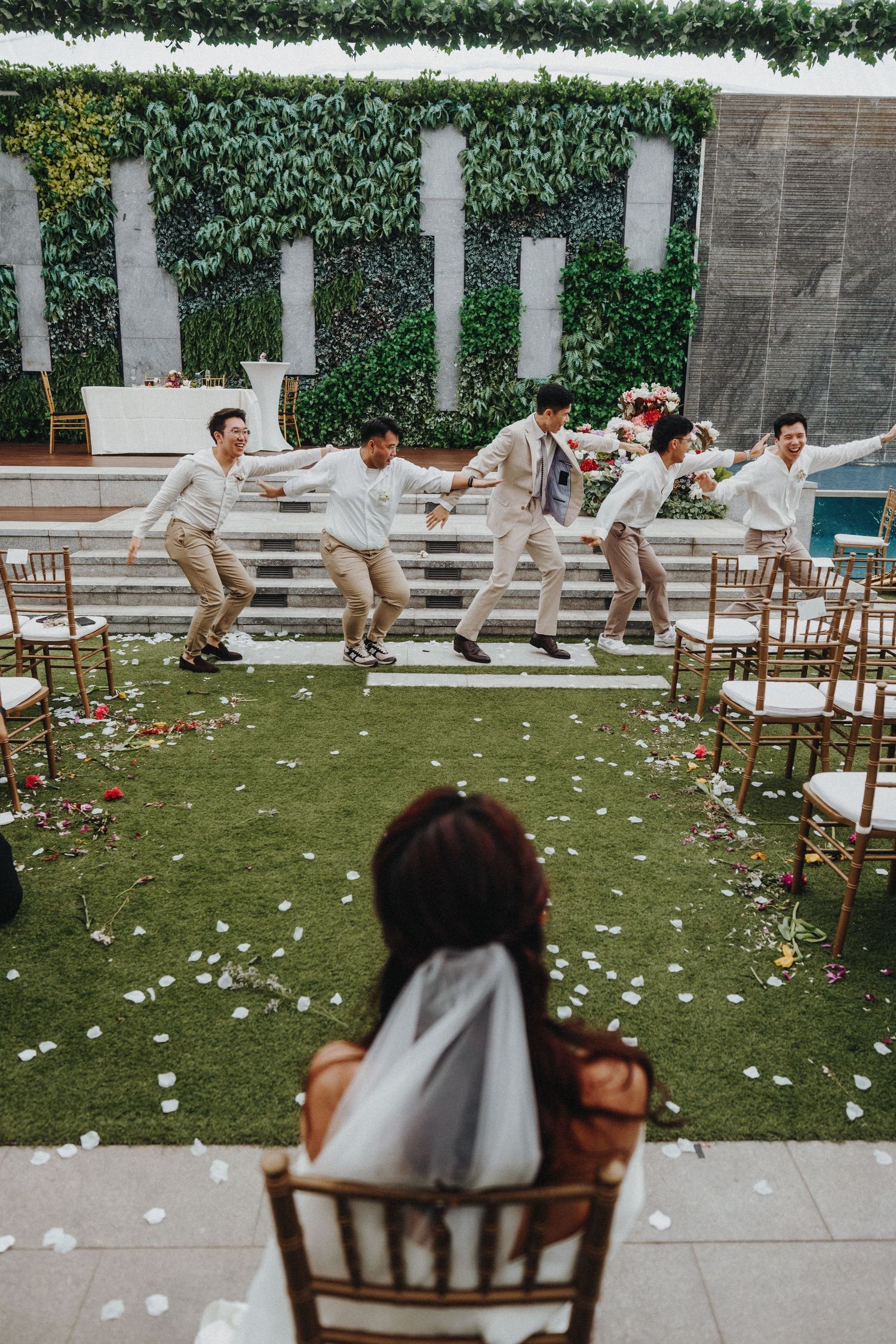 A joyful wedding party in beige suits dances in a line on outdoor steps while being watched from a wooden chair below.