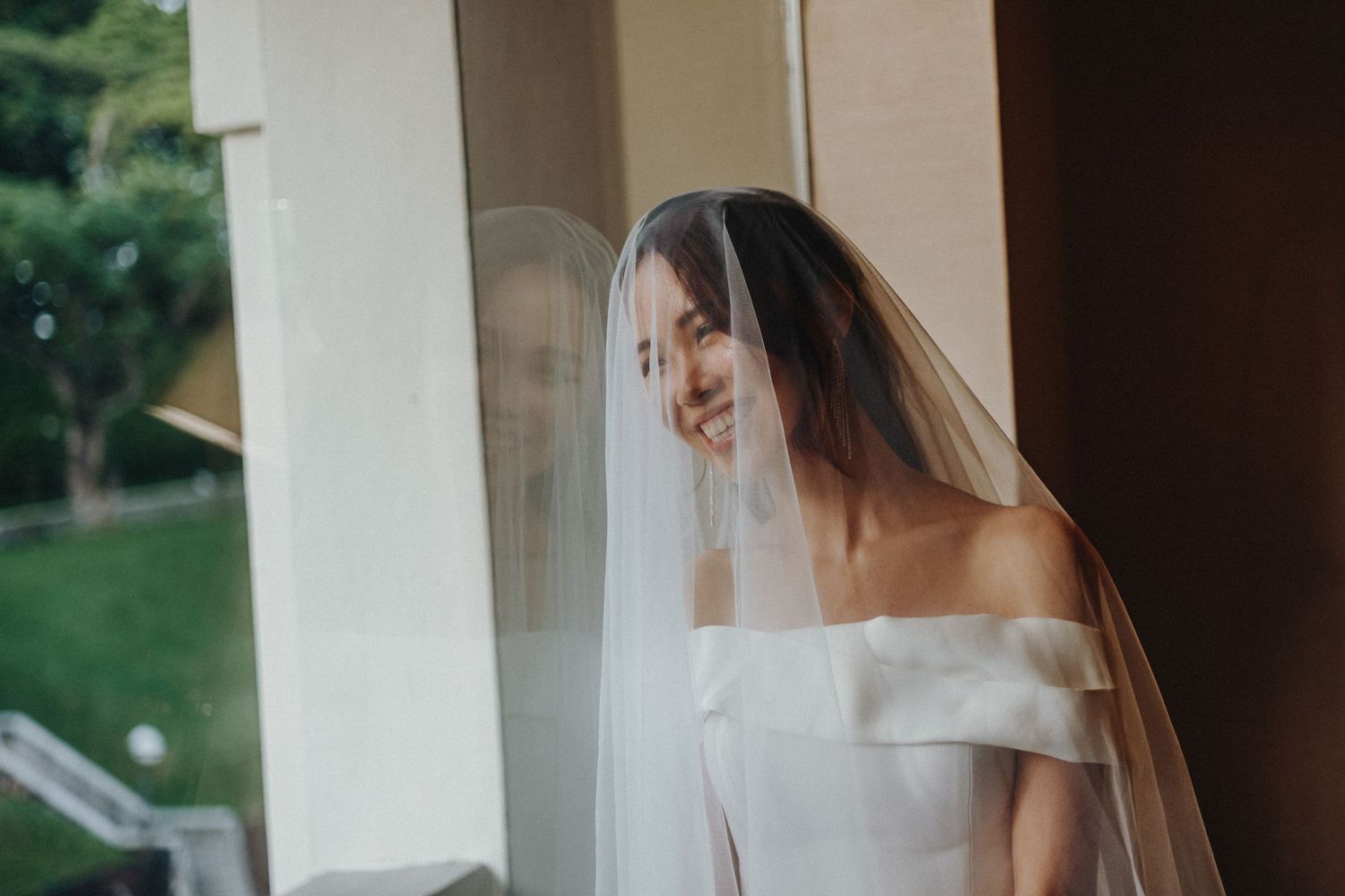 A bride in an elegant white gown stands by a window with flowing curtains in a romantic setting.