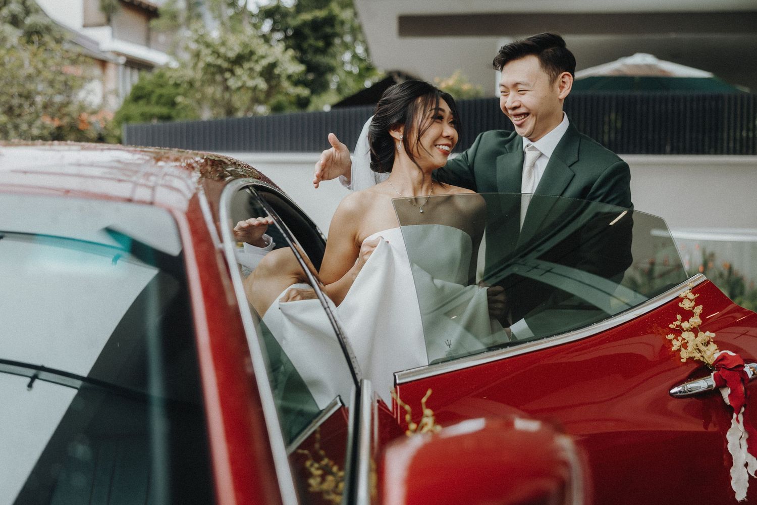 A joyful wedding couple sharing a laugh while standing by a decorated red car outdoors.