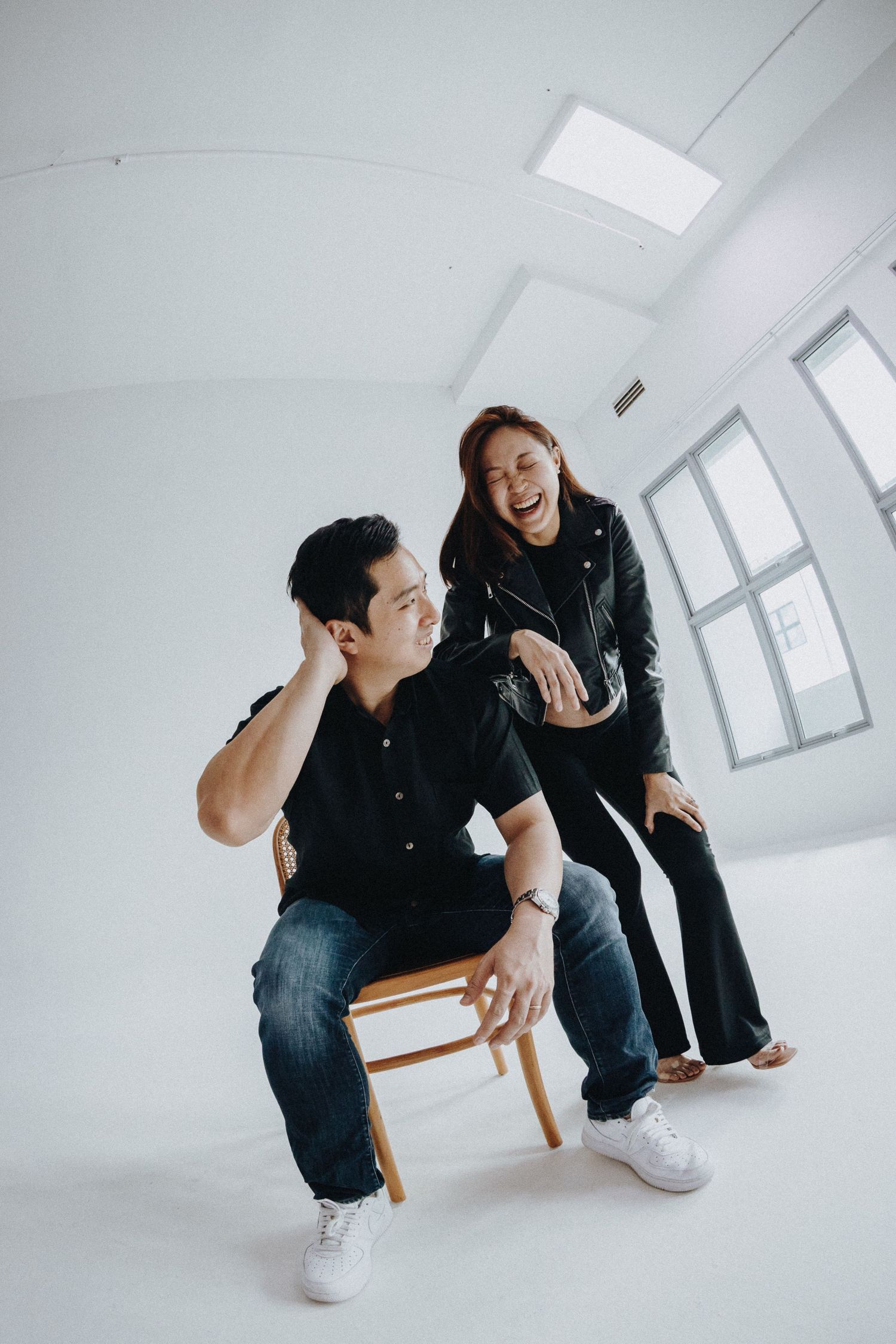A couple in dark clothing interacts playfully while sitting in a minimalist white room.