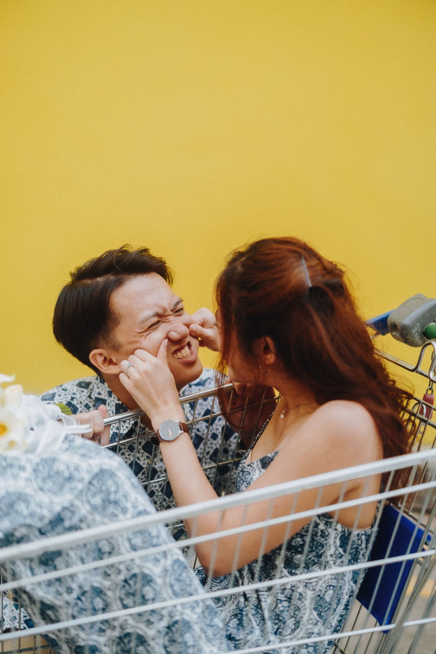 A candid laughing moment between a couple in a shopping cart against a yellow wall.