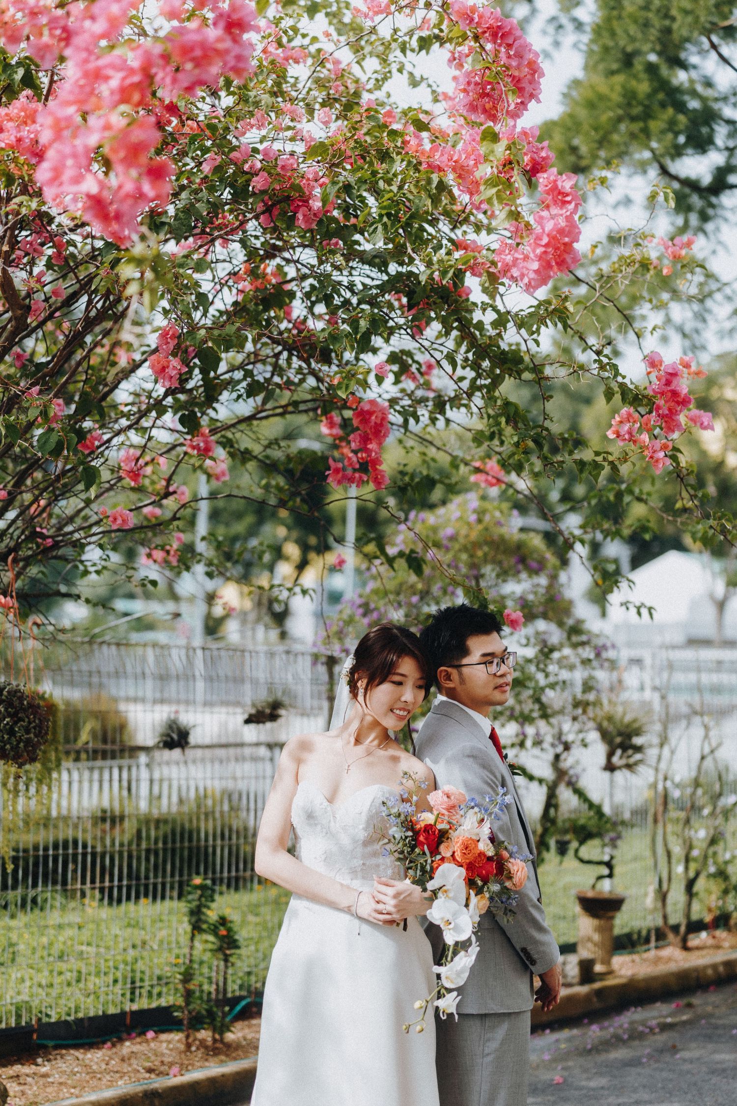 Wedding couple stands beneath vibrant pink bougainvillea flowers during outdoor photo session.