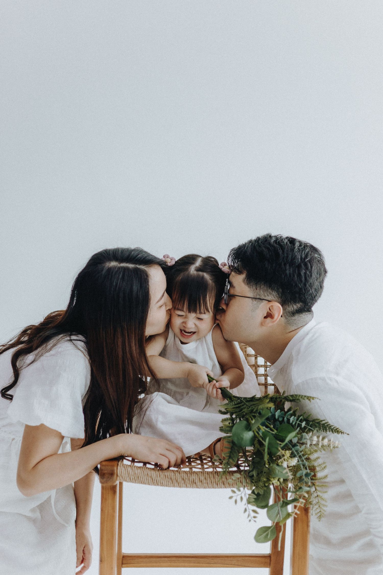 Intimate family moment captured in white outfits against light background with decorative plants.